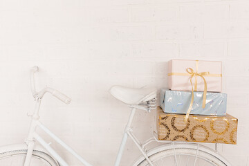 Original festive decor. White bicycle stands against background of white brick wall in back seat is pile of gift boxes. Wrapped in gift paper, tied with ribbons. Copy space.