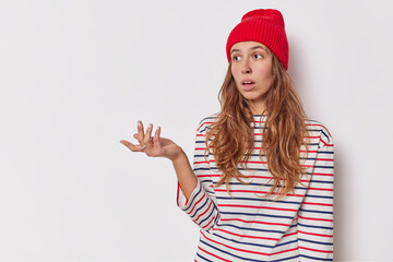 Confused indecisve woman wears red hat and casual striped jumper raises palm with hesitation faces...