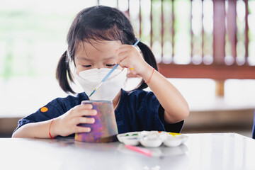 Cute girl is using her left hand to paint watercolor on pottery. Children wear white face mask to prevent spread coronavirus and PM 2.5 air pollution.