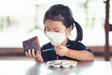 Child painting plant pot with watercolor. Kid wearing white face mask. Cute girl aged 4 years old.