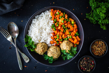 Boiled meatballs with white rice, carrots and peas on black, wooden table
