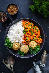 Boiled meatballs with white rice, carrots and peas on black, wooden table
