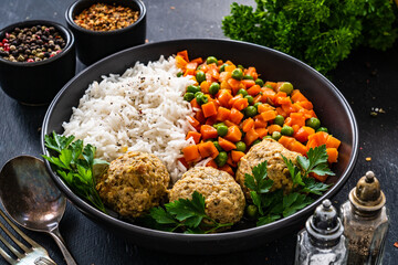 Boiled meatballs with white rice, carrots and peas on black, wooden table
