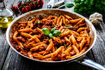 Penne with minced meat, tomato sauce, parmesan cheese, basil and vegetables served in frying pan on wooden table

