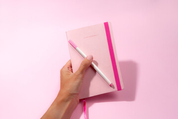 woman's hand holding a pink notebook with pen on pink background