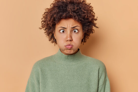 Photo of funny curly haired young woman pouts lips blows cheeks makes hilarious grimace holds breath crosses eyes dressed in casual jumper isolated over beige background tries to amuse someone