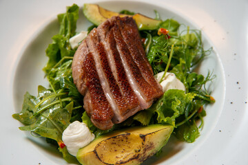 grilled duck steak with fresh vegetables and avocado on a white round plate on a wooden table close up