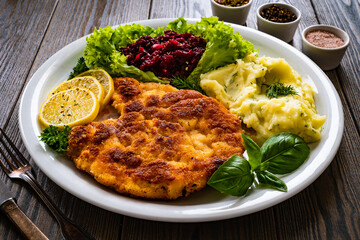 Breaded fried pork chop with potato puree, beetroots, lemon and fresh vegetables on wooden table
