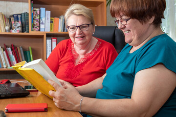 Two women are sitting at table, laughing, looking at book. Fun pastime. Generation of baby boomers. Selective focus.