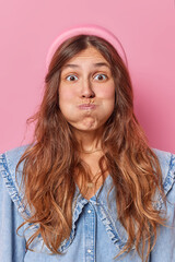 Vertical shot of funny long haired young woman puffes cheeks holds breath makes grimace wears...
