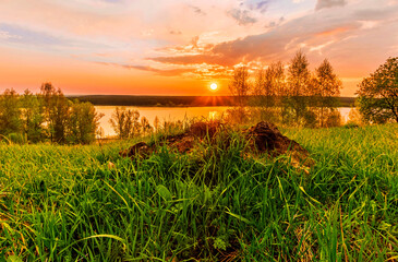 Scenic view at beautiful sunset on a shiny lake with old rough stone on the foreground, green grass, birch trees, golden sun rays, calm water ,nice cloudy sky on a background, spring landscape