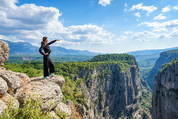 Side view of smiling female standing on edge of rocky cliff in highlands and pointing away on summer day