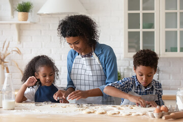 Fototapeta premium Cute little Black kids helping mom to bake cookies. Mother and sibling children cooking together, preparing dough for homemade biscuits at flour table, using cutters. Family household activities