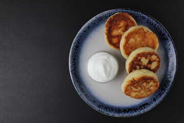 three cheesecakes lie on a blue plate with a sauce bowl of white sour cream sauce on a black background with space for text. Russian breakfast is traditional. Dietary proper nutrition. 