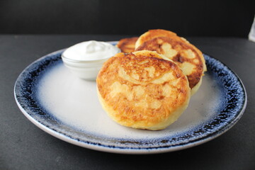 three cheesecakes lie on a blue plate with a sauce bowl of white sour cream sauce on a black background with space for text. Russian breakfast is traditional. Dietary proper nutrition. 