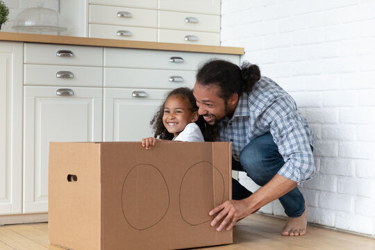 Joyful Black dad and excited little daughter girl playing driving toy car at home. Happy father dragging cardboard box with drawn automobile wheels and kid inside, Family fun, role games, fatherhood