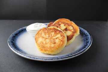 three cheesecakes lie on a blue plate with a sauce bowl of white sour cream sauce on a black background with space for text. Russian breakfast is traditional. Dietary proper nutrition. 
