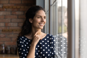 Relaxing and dreaming. Happy young hispanic female dreaming at home looking through big window watching beautiful good day outside. Smiling millennial lady rest on weekend lost in positive thoughts