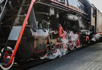 Steam comes under the wheels of the old locomative when departing from the station. Black iron train with red details