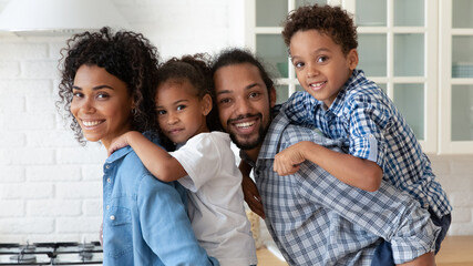Happy African American family couple holding little sibling kids, looking at camera, smiling for head shot portrait. Cheerful millennial parents piggybacking two children, having fun at home. Banner