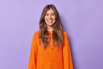 Fototapeta premium Portrait of pretty young European woman with long dark hair smiles pleasantly wears casual orange knitted sweater isolated over purple background shows white teeth. People and emotions concept