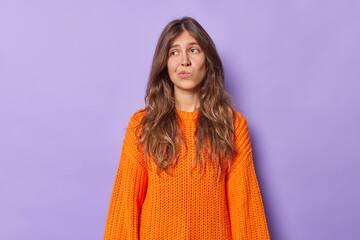 Photo of thoughtful long haired woman with bored expression hesitates about something purses lips wears knitted orange jumper isolated over purple background. People and face expressions concept