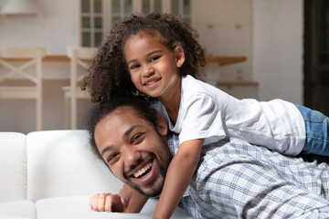 Fototapeta premium Cheerful African American daughter girl resting on happy dads back, looking at camera, smiling. Portrait of daddy lying on sofa at home, piggybacking child, enjoying leisure time, playing active games