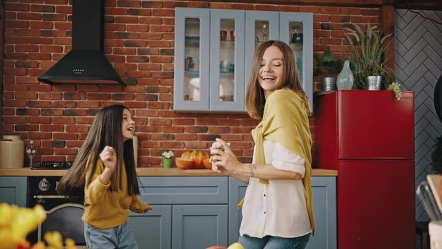 Young overjoyed mum and little daughter are laughing and dancing while cooking in modern kitchen at home. Slow motion
