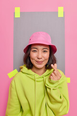 Vertical shot of pleased Asian girl makes korean like sign expresses love has cheerful expression wears panama and hoodie poses indoor blank copy space behind to place your advertising content
