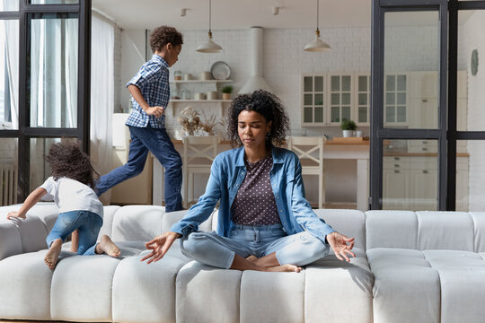 Calm African American Mom Meditating, Keeping Emotions Under Control, Sitting On Couch In Lotus Pose. Active Disobedient Sibling Children Running Around Peaceful Mother. Kids At Home Concept