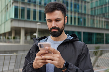 Serious handsome guy holds mobile phone looks attentively at screen sends message to friends in social networks downloads maps application poses near modern building in city wears anorak smartwatch © WHstudio Leushin N
