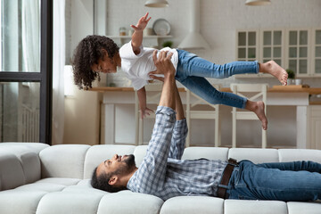 Happy strong African dad lifting little daughter girl up in air, holding kid with flying open...