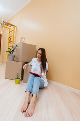 Fototapeta na wymiar A young woman is sitting on the floor in front of the boxes and picks up a new design for her apartment on a tablet after moving. Housewarming, delivery and cargo transportation.
