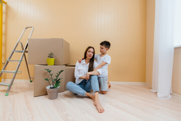 Fototapeta na wymiar A young woman with her son is sitting in front of the boxes and rejoicing at the housewarming after moving in. Housewarming, delivery and freight transportation, purchase of real estate.