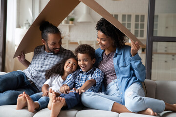 Happy joyful Black millennial couple and little sibling kids having fun, playing together on couch, holding carton roof, symbol of house, safety insurance. Homeowners family enjoying new home