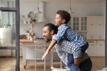 Happy African American father and son enjoying active games at home, going wild. Daddy carrying boy on back, piggybacking kid, playing airplane, running, exercising at home. Family having fun