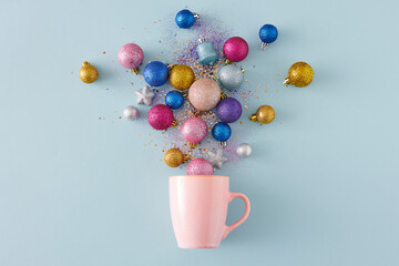 Top view of Christmas tree balls and sequins flying out of pink cup on isolated pastel blue...