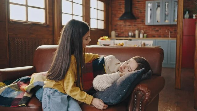 Young mother fell asleep with book on couch in kitchen of country house. Little daughter coming up to her, covering mother with plaid and hugging