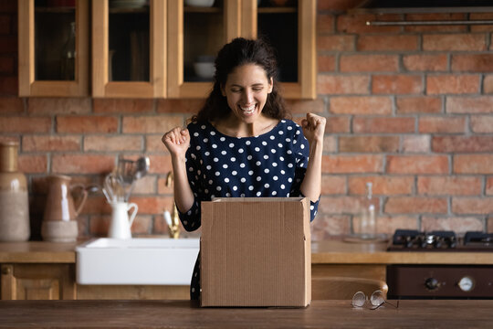 Shipment For Web Shopper. Overjoyed Latin Female Customer Open Delivery Box At Kitchen Table Excited With Internet Order Received From Ecommerce Website. Happy Young Lady Unpack Postal Parcel Get Gift