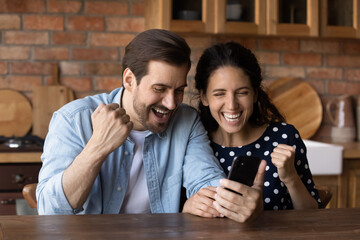 Great thing. Excited young married couple sit at kitchen hold smartphone yell yes celebrate getting...