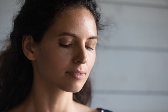 Tender face. Close up of calm peaceful millennial hispanic woman with beautiful fresh clear skin looking down breathing air meditating with closed eyes. Grey boarded fence is on background. Copy space