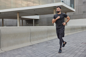 Determined bearded adult man in activewear jogs in city street covers long distance enjoys morning...