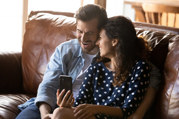 Funny thing. Overjoyed young married couple cuddle on sofa watch cute comedy hilarious video clips at social media. Laughing millennial boyfriend girlfriend having fun enjoy amusing photos on cell