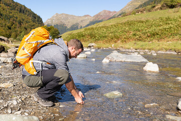 Male traveler with a orange backpack washing his hands in a mountain river. Aventure travel.  - Powered by Adobe