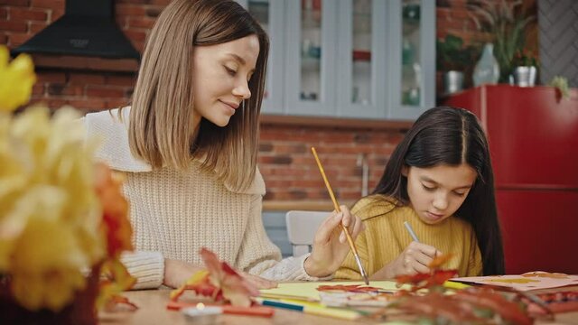 Young mother and preteen daughter smiling, drawing leaves with brush and orange watercolor paint, sitting at table in kitchen. Close up