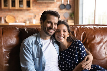 Newlyweds. Portrait of cheerful bonding spouses husband wife hug on sofa look at camera with healthy toothy smiles. Happy young couple in love homeowners renters tenants of new house enjoy family life