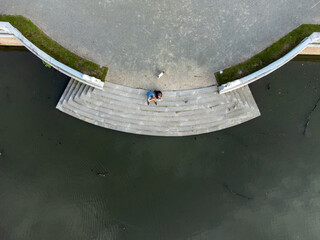 People sitting on steps by the water with a small white dog behind them. Drone aerial shot at Hof ter Linde in Edegem