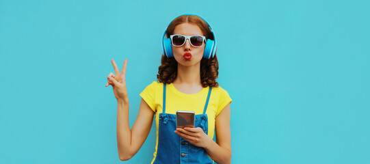 Fashionable portrait of stylish young woman in headphones listening to music with smartphone on blue background
