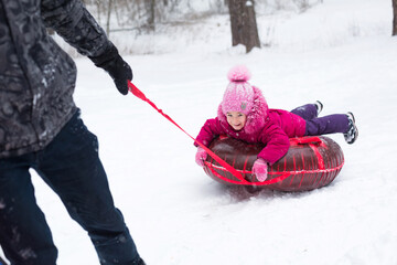 Dad rides his daughter on a tube  from a slide in winter in a snowfall. Tubing, winter sports outdoor and family entertainment. Warm clothes, knitted hat with a pompom. Help and support, family values