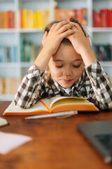 Close-up vertical shot of exhausted primary child school girl tired from studying holding head head with hands while reading book sitting at desk with workbook. Child schoolgirl doing homework at home